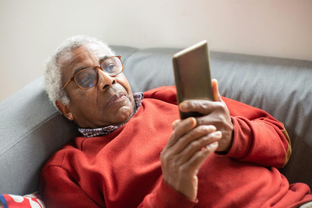 Elderly man relaxing indoors using a smartphone, wearing glasses and a cozy sweater.